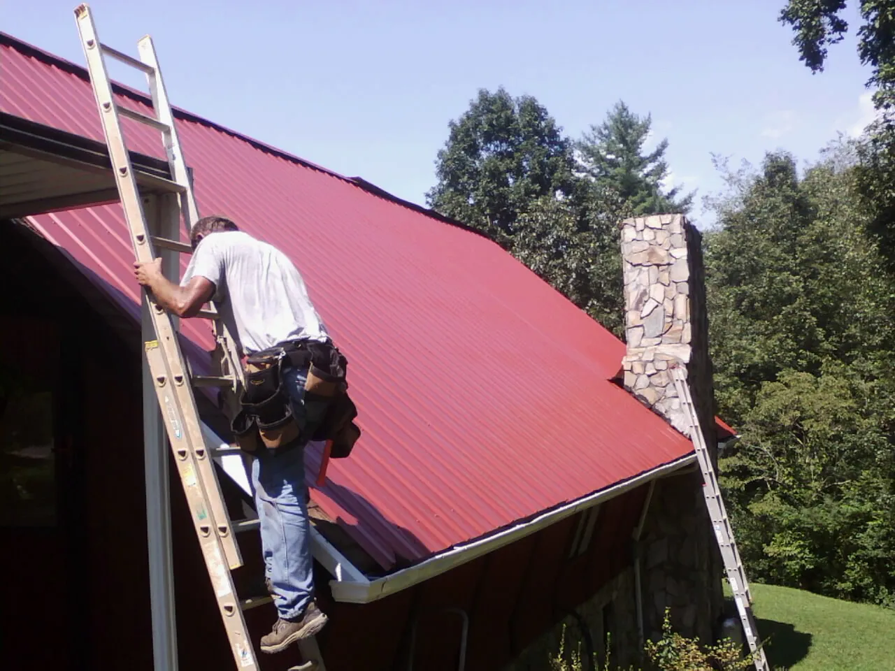 Roofer on a ladder beside a red metal roof and stone chimney — Soffit & Fascia Repair by Roofing Brothers in Axtell
